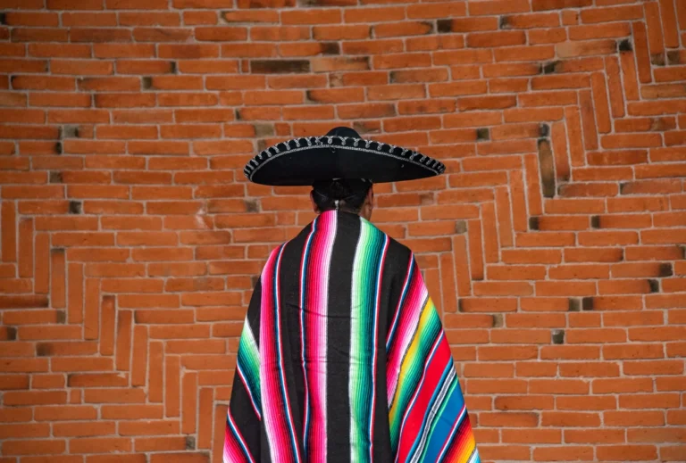 A person in colorful Mexican attire and sombrero standing against a brick wall.