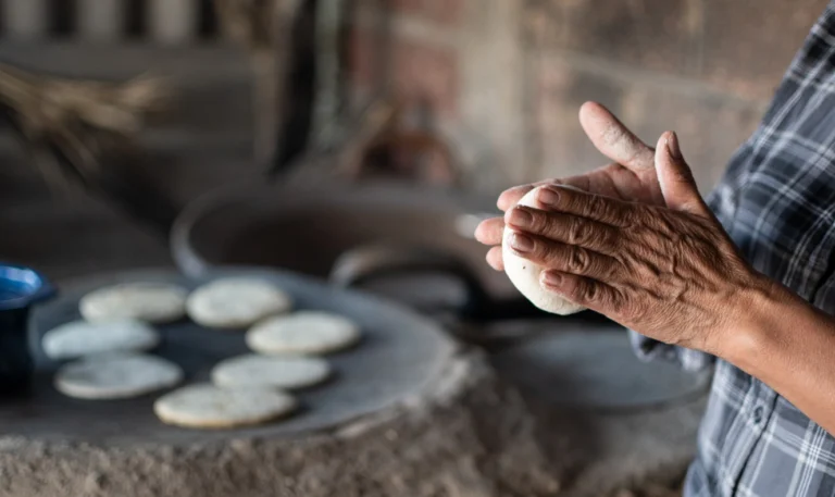 A person shaping dough with hands near cooked tortillas on a surface.