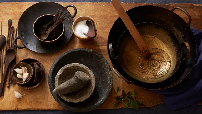 A variety of kitchen tools including wooden spoons, bowls, and a mortar on a wooden surface.