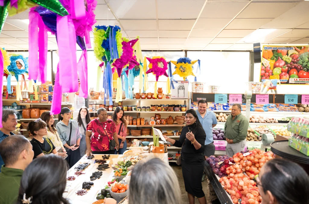 A group of people listening to a guide near a display of fresh produce and artisanal goods in a brightly decorated market.