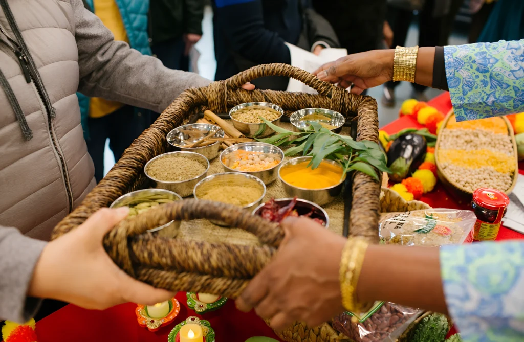 Two pairs of hands passing a woven tray containing small metal bowls filled with various spices, lentils, and fresh herbs.